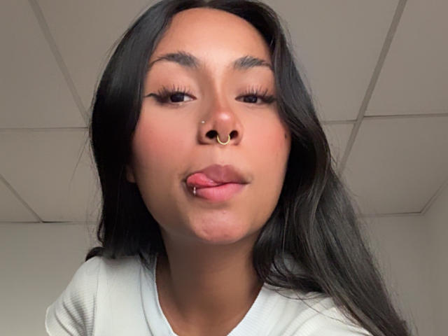 AnaRome has long black hair, a nose ring, and is sticking out her tongue with her lips slightly puckered. She's wearing a white shirt and has long eyelashes.