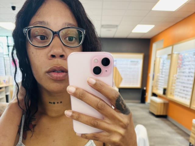 LexxxBright with glasses, wavy dark hair, and a choker, holding a pink iPhone with three cameras, taking a selfie, showing a tattoo on her right hand.