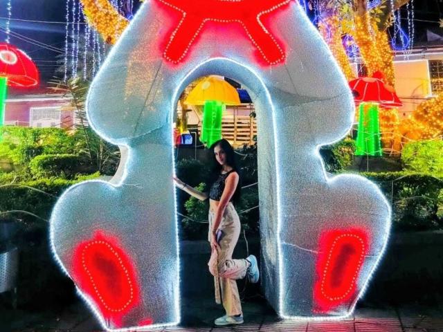 LilithDesire stands in front of a glowing, heart-shaped LED arch, wearing a black top and beige pants, with a small white bag in her right hand.