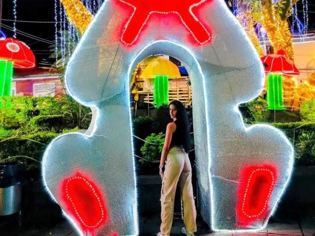 LilithDesire stands in front of a glowing, large, neon blue 'T' with red accents, wearing a black top and beige pants, showing her round ass.