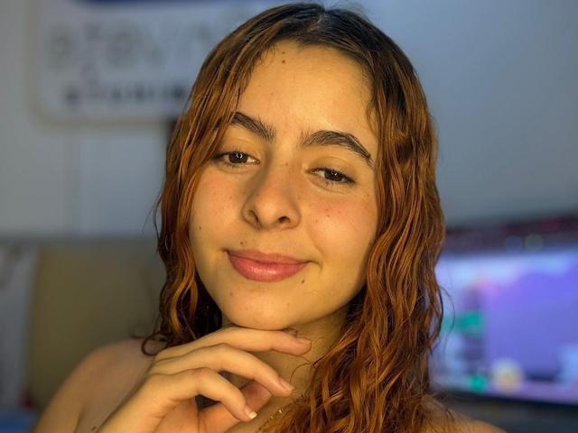 AdeleiineFoster with wavy brown hair, light skin, small nose, and subtle smile, touching her chin, in a blurry room with a TV in the background.
