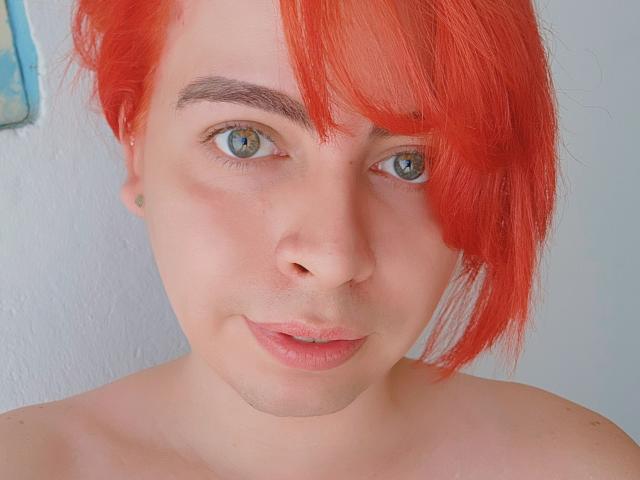 Close-up of TsFoxCandy with bright red hair, green eyes, and fair skin. The hair is slightly messy, covering the right side of the face. TsFoxCandy has a small, subtle mustache and a neutral, slightly smiling expression. The eyebrows are dark and well-groomed. The background is a simple, light blue wall with a small, partially visible painting on the left. The image is sharply focused on TsFoxCandy's face, highlighting the natural texture of their skin and the vibrant color of their hair.