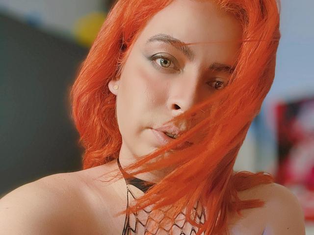 TsFoxCandy with bright red hair, green eyes, and a nose ring, has a serious expression, a black choker, and a wisp of hair in her mouth.