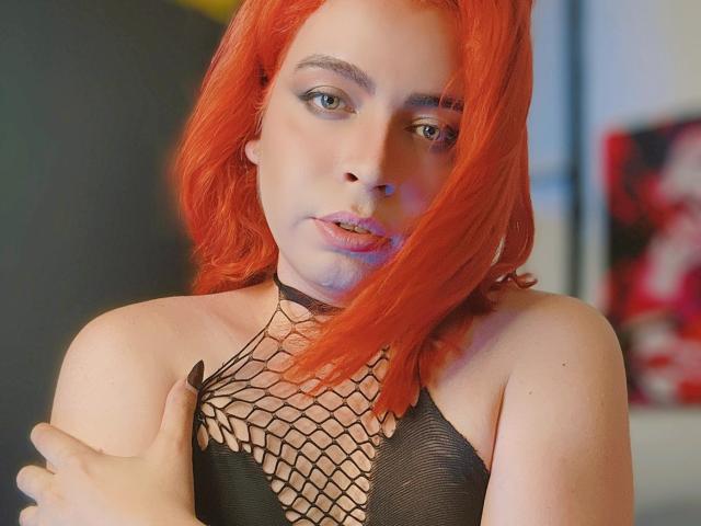 TsFoxCandy with bright red hair, wearing a black fishnet top, showing her small tits, holding her arm, looking at the camera with blue eyes and pink lipstick.