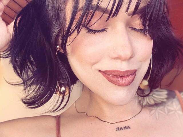 HannaBorbon has black shoulder-length hair, closed eyes, and dark lipstick. She's wearing gold earrings and a thin necklace, with a tattoo above her right breast.