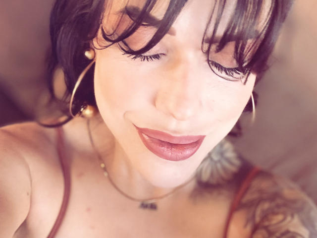 HannaBorbon's face, eyes closed, dark lipstick, black bangs, hoop earrings, necklace, tattoo on right shoulder, soft light, smiling, upper chest visible, no clothes shown.