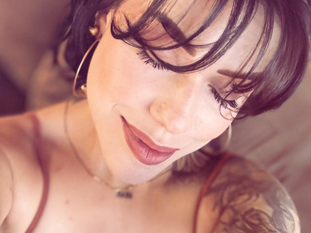 HannaBorbon, con i capelli neri corti, gli occhi chiusi e il rossetto rosso, indossa orecchini a cerchio e una canotta rossa, mostrando un tatuaggio sulla spalla destra, e sorride dolcemente..
