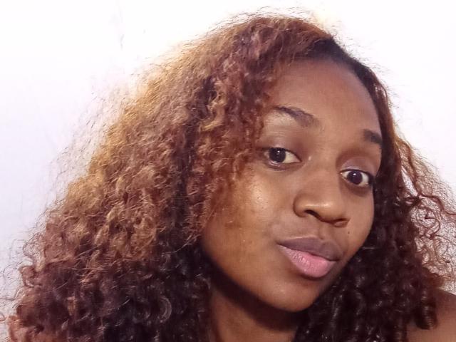 LindahSexy has curly brown hair with lighter tips, brown eyes, and pink lips. She's looking slightly to the side with a subtle smile. Her skin is dark brown.
