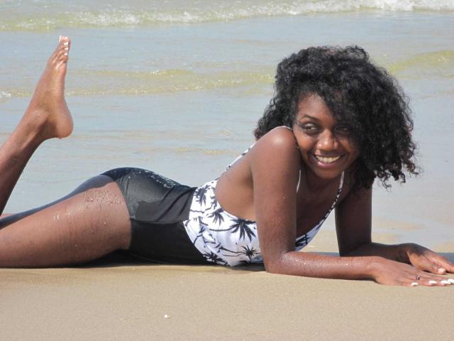 LindahSexy, lying on sandy beach, wearing a black and white floral one-piece, showing her ass, smiling with curly black hair, and water droplets on her dark skin.