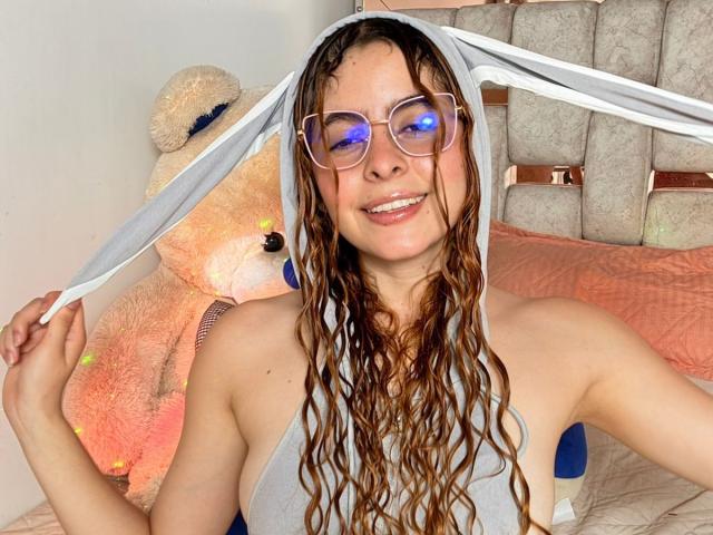 AdeleiineFoster with wavy brown hair, wearing glasses with blue light, grey one-shoulder top, and bunny ears, smiling at the camera, next to a large stuffed teddy bear.