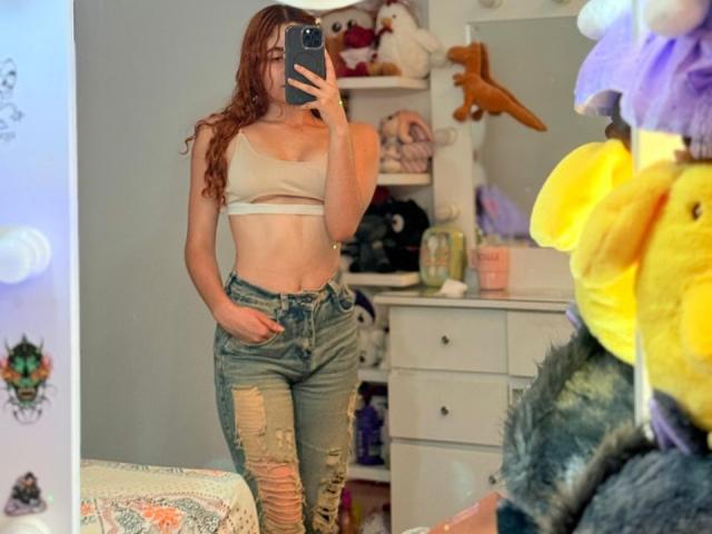AdeleiineFoster with wet red hair, wearing a beige bra and ripped blue jeans, taking a mirror selfie, hand in pocket, showing her small tits and slim ass.