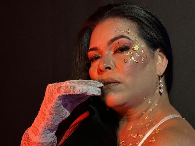 JesikaSweety with dark hair, wearing a white strap, has glitter on her cheek and neck, and a lace glove on her right hand. She touches her cheek with the glove, looking to the side. She has a silver earring in her right ear. The lighting is red and warm, highlighting her face and glitter. Her makeup is bold, with dark eyeshadow. The image is close-up, focusing on her face and upper body.