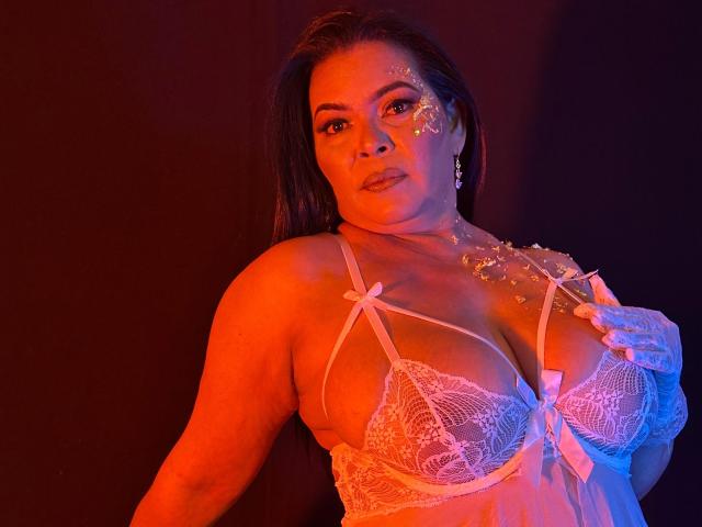 JesikaSweety, a plus-sized woman with long dark hair, stands against a dark red-orange background. She wears a sheer white lace bra with pink ribbon accents, highlighting her large breasts. Her right hand touches the bra's strap, and she has glitter on her left cheek. She has a serious expression and wears small hoop earrings. The lighting casts a warm, orange-red glow on her skin. The image has a sensual, provocative feel.