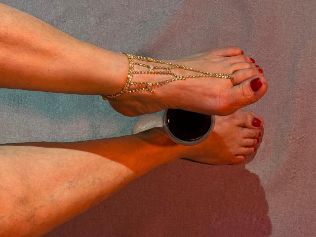 Two feet with red-painted toenails, JesikaSweety's right foot is above her left, adorned with a gold chain anklet. Feet are slightly dirty, with a small shadow on the grayish carpet. The anklet has multiple thin gold chains, each chain connecting to her ankle. JesikaSweety's right heel is over a small black cup. The image is sharply focused, emphasizing the texture of the anklet and the paint on her toenails.