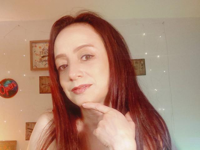 CatalinaMonyque with red hair, light skin, and subtle makeup, gently touching her chin with her finger, smiling slightly. She's topless, showing her bare shoulders.