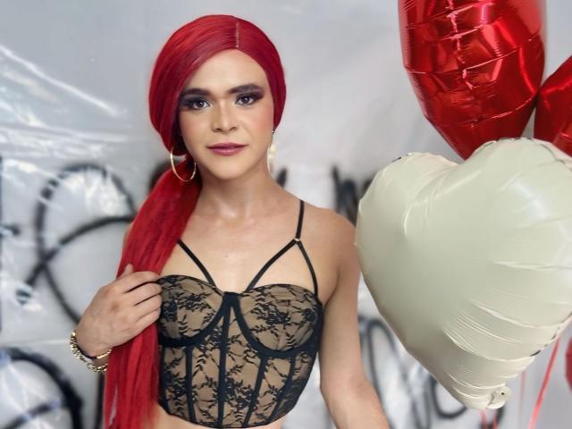 Shaina Monroe met rood haar, die een zwarte kanten lingerie top draagt, witte en rode ballonnen vasthoudt, oorringen en een armband, waarbij haar borsten zichtbaar zijn, en die naar de camera kijkt..