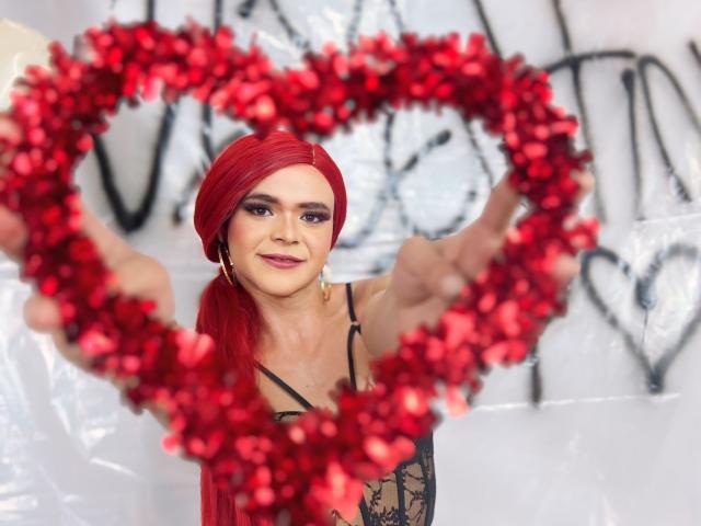 Shaina Monroe met rood haar, die een rode, hartvormige bloemenkrans vasthoudt, draagt een zwarte kanten jurk, glimlacht, heeft haar armen gespreid, haar borsten zijn zichtbaar, de achtergrond is wazig, de kleuren zijn rood en zwart..