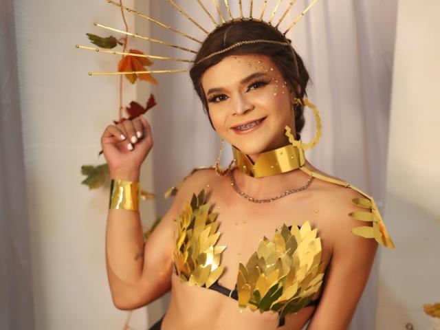 Shaina Monroe draagt een beha met gouden bladeren, een gouden hoofdtooi en armbanden, en houdt herfstbladeren vast, terwijl ze glimlacht. Haar borsten zijn bedekt met gouden bladeren, en ze draagt een ketting..