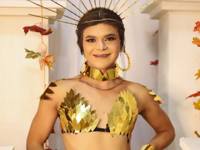 Shaina Monroe con reggiseno e copricapo dorati, e collana a catena, sorride con le mani sui fianchi, indossando bracciali dorati e orecchini a cerchio dorati, appare incredibilmente sexy..