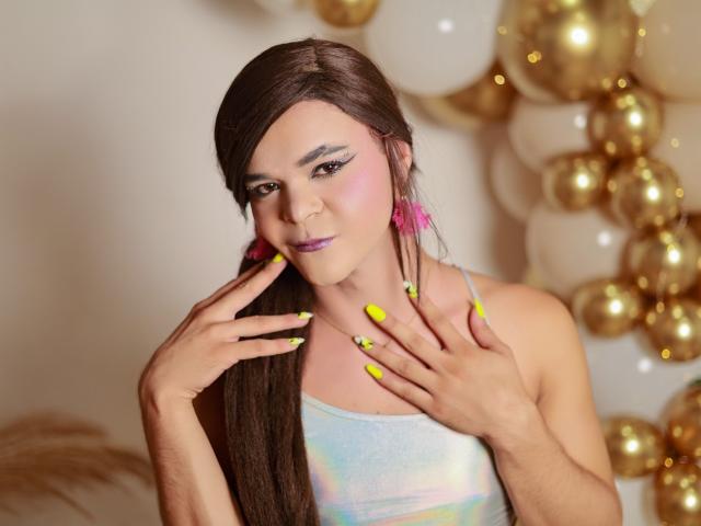 Shaina Monroe met lang bruin haar, roze oorbellen, gele nagels en een kleurrijke top, die haar gezicht aanraakt en de camera aankijkt met een lichte glimlach..