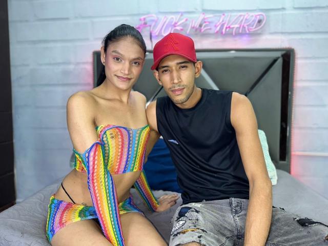 UnitedSou with rainbow striped bikini, showing her tits, sits next to UnitedSou in a red cap, black sleeveless shirt, and ripped jeans, both smiling on a bed.