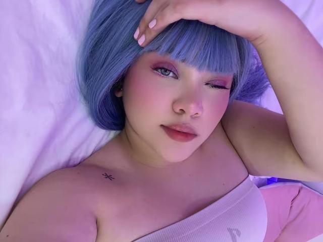 AlayaWillow with blue hair, winking, lying on a bed, hand on head, wearing a white strapless top, pink eye makeup, small tattoo on right shoulder, pink lips.