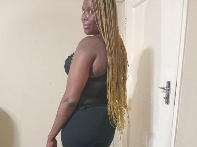 CaramelBootyX has dark skin, long blonde braids, black lace bra, and black pants, standing with a side glance, showing her curvy ass and small tits.