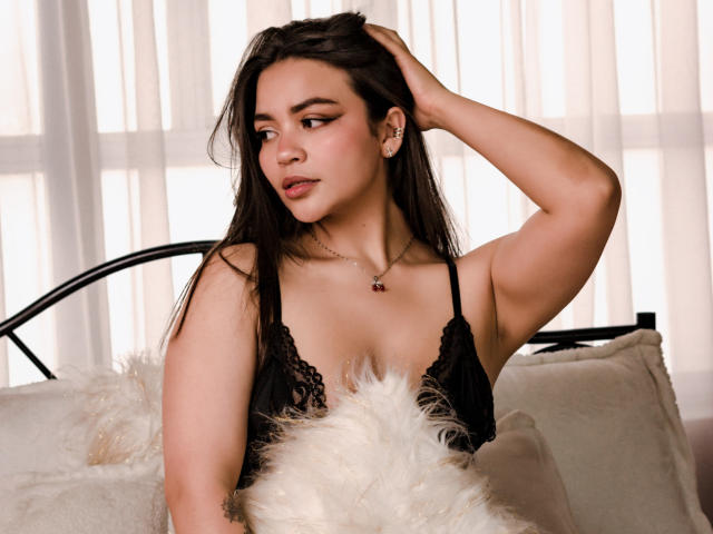 KimDevinne, con cabello largo y oscuro, una blusa de encaje negro y un collar, con la mano en el cabello, mirando hacia un lado, un almohadón blanco esponjoso, luz suave, linda, sexy, piel cálida, maquillaje sutil..