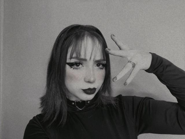 DarkCourtney with dark lipstick, black eyeliner, and a choker, wearing a black long-sleeve shirt, making a peace sign with a ringed finger, in a black-and-white photo.