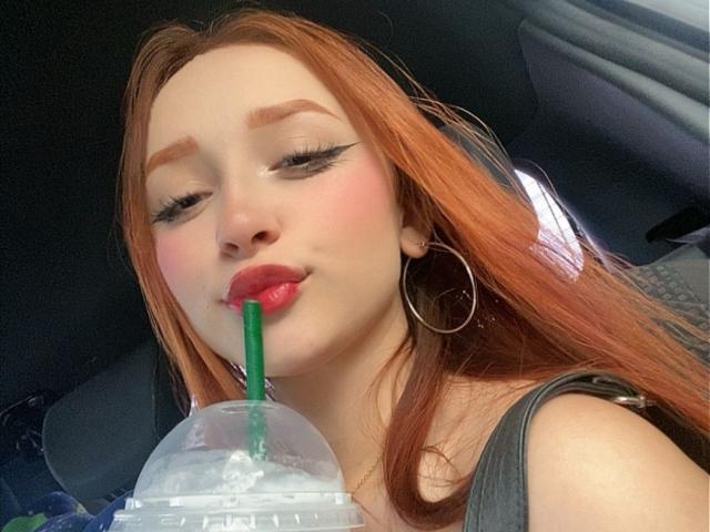 DarkCourtney with red hair, green straw in clear cup, pink lips, hoop earrings, and eyeliner, sucking on straw in car. Her tits are barely visible under gray tank top.