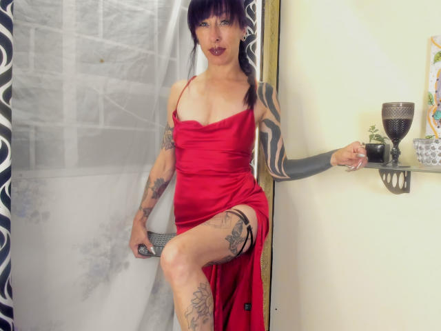 GrethelMulher with tattoos, wearing a red dress, sitting with one leg up, holding a remote, and looking at the camera with dark lipstick, in a room with a shelf.
