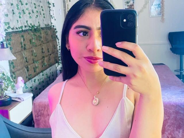 ZoeRedds with pink lipstick, holding a black phone, wearing a white tank top, necklace with a pink gem, taking a mirror selfie, showing her tits.