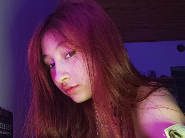 LemyRose met lang rood haar, een neusring en een ketting, kijkt in de camera met een lichte frons, onder paars en roze verlichting, waarbij haar borsten zichtbaar zijn..