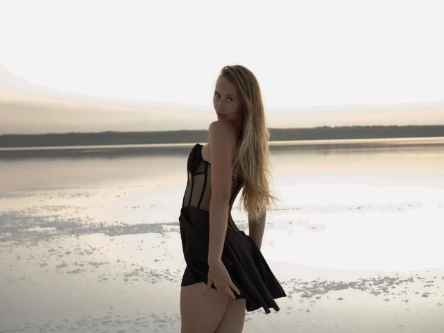 MovieStarI with long blonde hair, wearing a black, sheer, strapless dress, showing her ass, standing by a calm, reflective water body, looking over her shoulder.