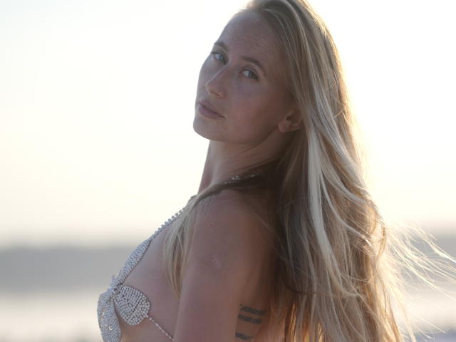 MovieStarI hat langes, blondes Haar, ein silbernes Bikini-Oberteil und ein Tattoo auf ihrem rechten Arm. Sie blickt über ihre Schulter und lächelt leicht. Das Sonnenlicht scheint von hinten auf sie..