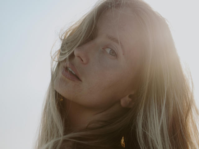 A atriz MovieStarI tem cabelo loiro, pele clara e está olhando para cima com os lábios entreabertos, com o sol refletido nos olhos e um leve vento em seus cabelos. Seus seios não estão visíveis..
