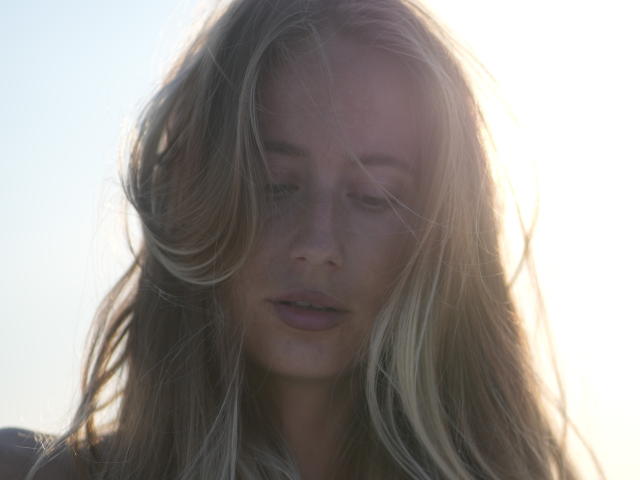 MovieStarI a de longs cheveux blonds ondulés qui lui cachent le visage, avec des lèvres légèrement entrouvertes, illuminées par le soleil, et une expression douce et sereine, tournée vers le bas. Son visage est légèrement flouté par la lumière..