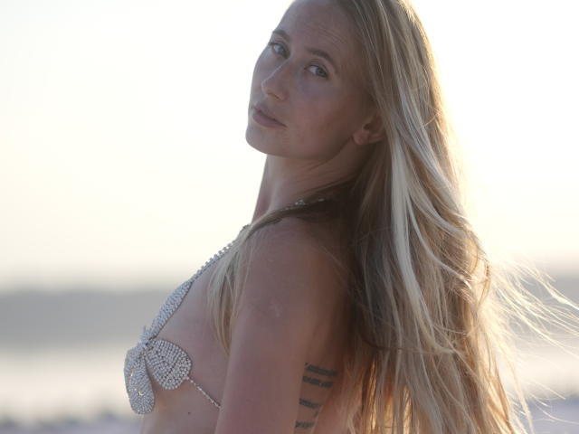 Ein Filmstar mit langen blonden Haaren, der ein glitzerndes BH-Top trägt und mit einem leichten Lächeln zurückblickt. Auf der rechten Seite der Brust ist ein Tattoo sichtbar. Weiches Sonnenuntergangslicht..