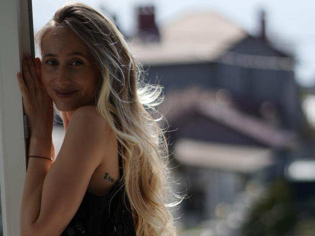 Ein Filmstar mit langen, blonden Haaren, lächelnd, an ein Fenster gelehnt, in einem schwarzen Spitzenkleid, kleine Tätowierung auf der linken Seite, Sonnenlicht in den Haaren, unscharfer Hintergrund, sichtbare Brüste, sanftes Lächeln..
