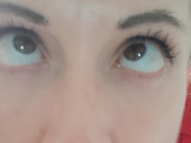 Close-up of LunaBrice's eyes looking upward. Her brown eyes are wide with light blue irises, framed by thick, dark eyebrows. She has long, black eyelashes on both eyes. The skin is pale with a slight pinkish hue near the eyes. The image is slightly blurred, emphasizing the eyes and eyelashes. The red background is partially visible.