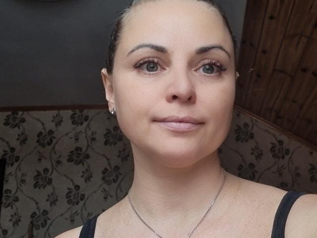 AlinaBurleck has fair skin, green eyes, and dark brown hair in a bun. She's wearing a black tank top and a thin necklace. Her small tits are visible.