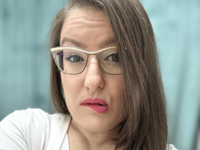BadAngel met een serieuze uitdrukking, die een bril draagt, roze lippenstift en een wit shirt, met bruin, recht haar dat één oog bedekt, en die lichtelijk opzij kijkt..