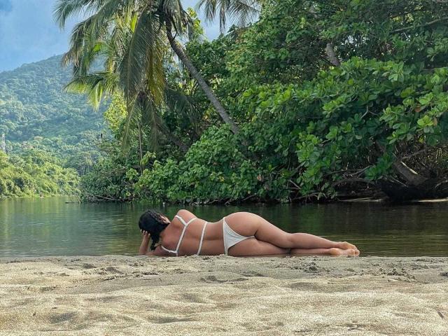 LucyySky è sdraiata su una spiaggia sabbiosa, rivolta di spalle, indossando un bikini bianco che mette in mostra i glutei e la schiena, con i capelli neri sciolti, vicino a un fiume calmo, circondato da alberi verdi..