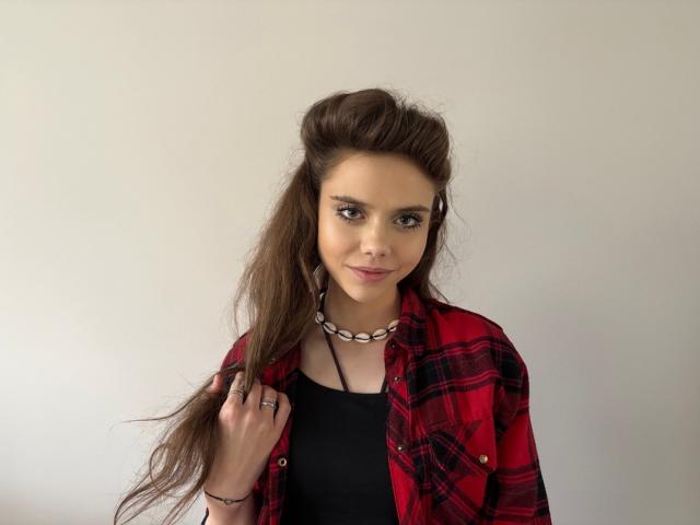 LilyGentle with long brown hair in a ponytail, wearing a red plaid shirt over a black top, necklace, and ring, smiling softly at the camera.