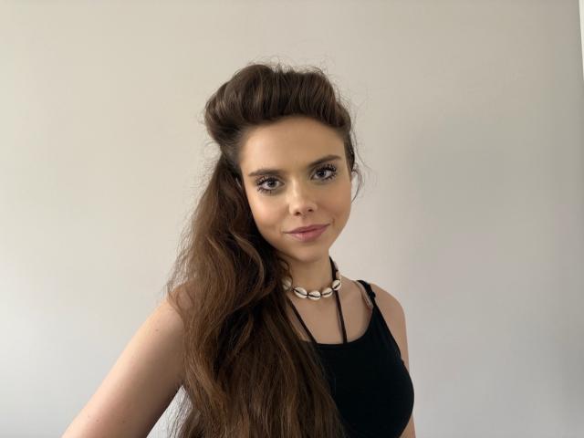 LilyGentle has long brown hair, wearing a black tank top, and a white chunky necklace, with a slight smile and relaxed expression. She has small tits and fair skin.