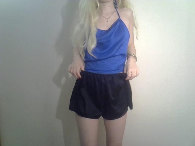 DitaVira stands against a plain wall wearing a bright blue halter-top and black shorts. Her long, wavy blonde hair cascades over her shoulders. She has a slim build with fair skin. Her hands are pulling down the shorts slightly. She wears a simple necklace and a bracelet on her left wrist. The image is straightforward with no additional elements or distractions. The focus is on her casual, summery outfit and her relaxed posture.