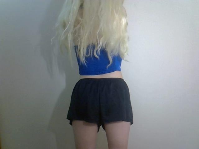 Blonde DitaVira with wavy hair in a blue top and black shorts, showing her ass, standing against a plain wall. Her back is to the camera.