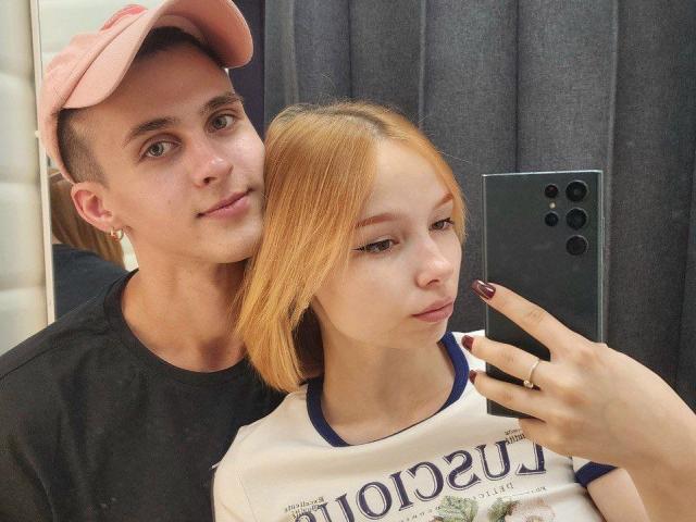 A selfie photograph featuring GuessDuality, a young man with light skin, short brown hair, and a pink cap, standing behind GuessDuality, a young woman with light skin and straight orange hair. GuessDuality wears a black t-shirt, while GuessDuality wears a white t-shirt with "LOUNA" text. GuessDuality holds a black smartphone with three cameras, taking the selfie. The woman's nails are painted dark red. The man's left ear has a small earring. The image is clear with a focus on the subjects' faces and the phone.