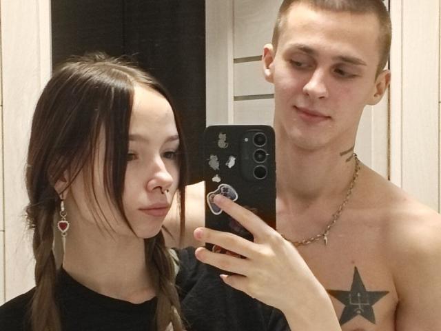 Selfie of GuessDuality and GuessDuality. GuessDuality on the left has long brown hair, a nose ring, and heart-shaped earrings. GuessDuality on the right is shirtless, has a shaved head, a star tattoo on the chest, and a gold chain necklace. GuessDuality on the right holds a black phone with a sticker on the back, taking the selfie. The photo is in a bathroom with white tiles. Both have neutral expressions.