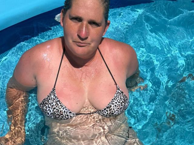 A chubby woman with wet skin and blonde hair wears a leopard-print bikini, her large tits visible above the blue water. She looks at the camera with a slight smile.